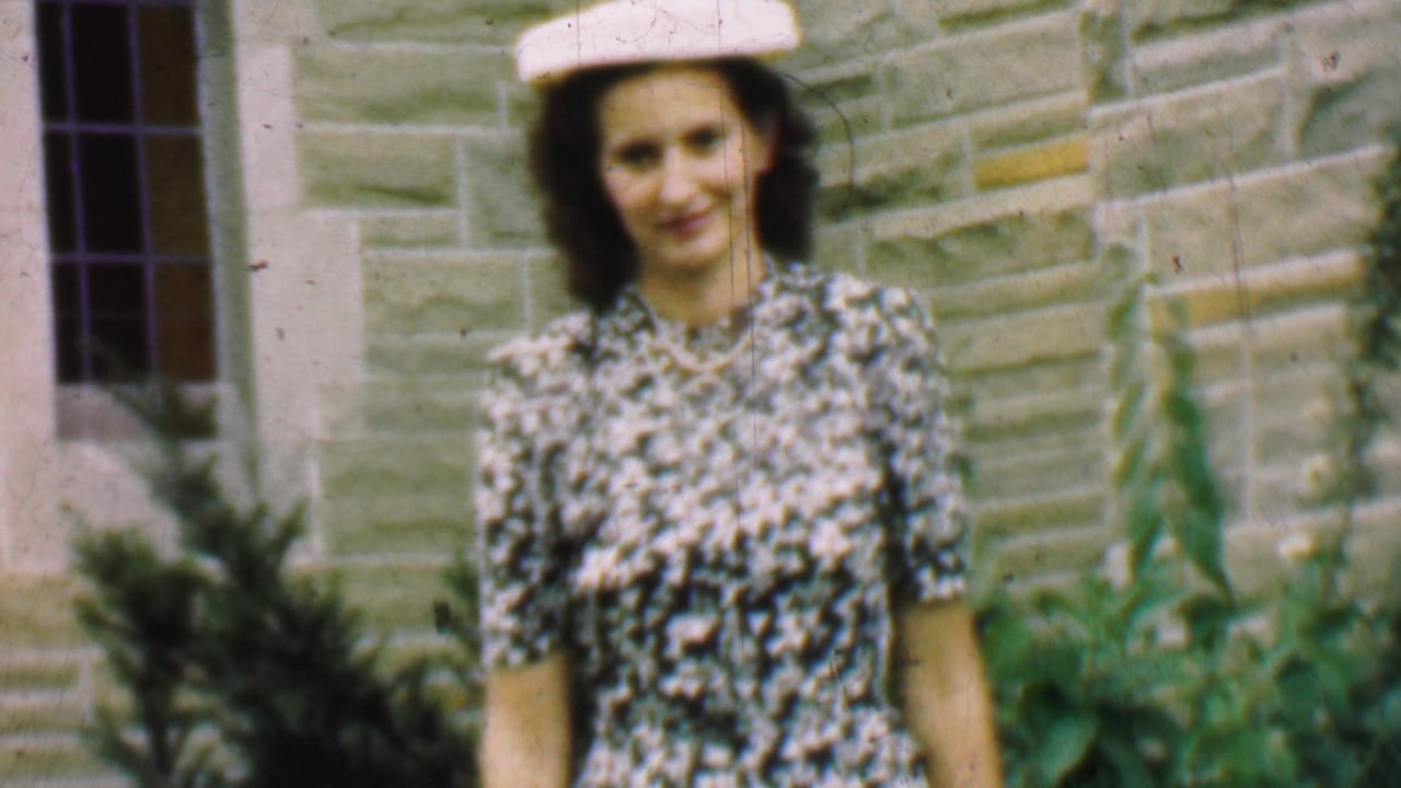 In metadata-supported 1950 Detroit home-movie footage, a woman in a white hat and black-and-white patterned dress stands outside a stone building with shrubs and a dark-paned window nearby.