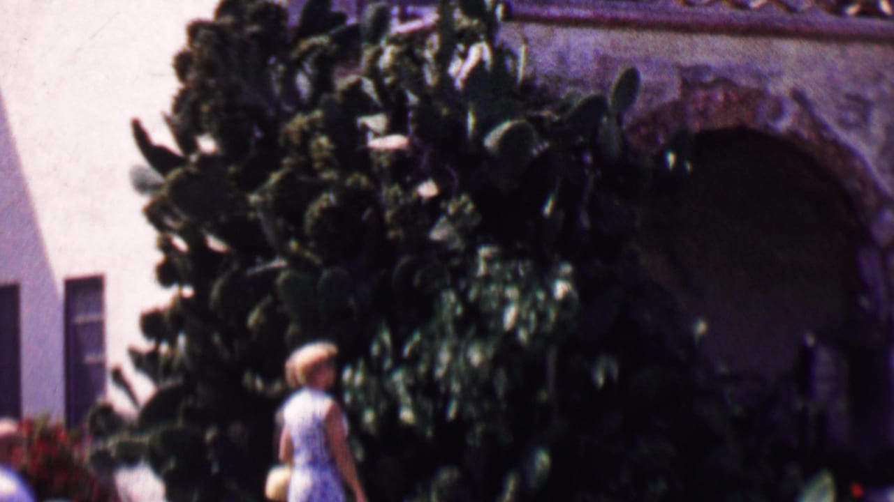 1955 Solvang home-movie frame showing a woman in a light dress walking beside dense cactus plantings in front of an arched stucco building.