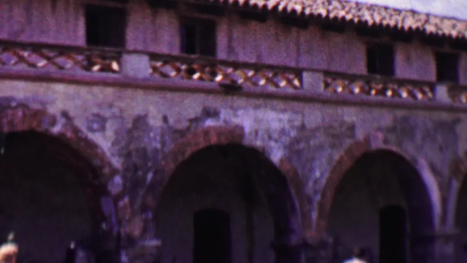 1955 Solvang home-movie frame showing a long arched facade with a tiled roofline and partial pedestrians along the lower edge.