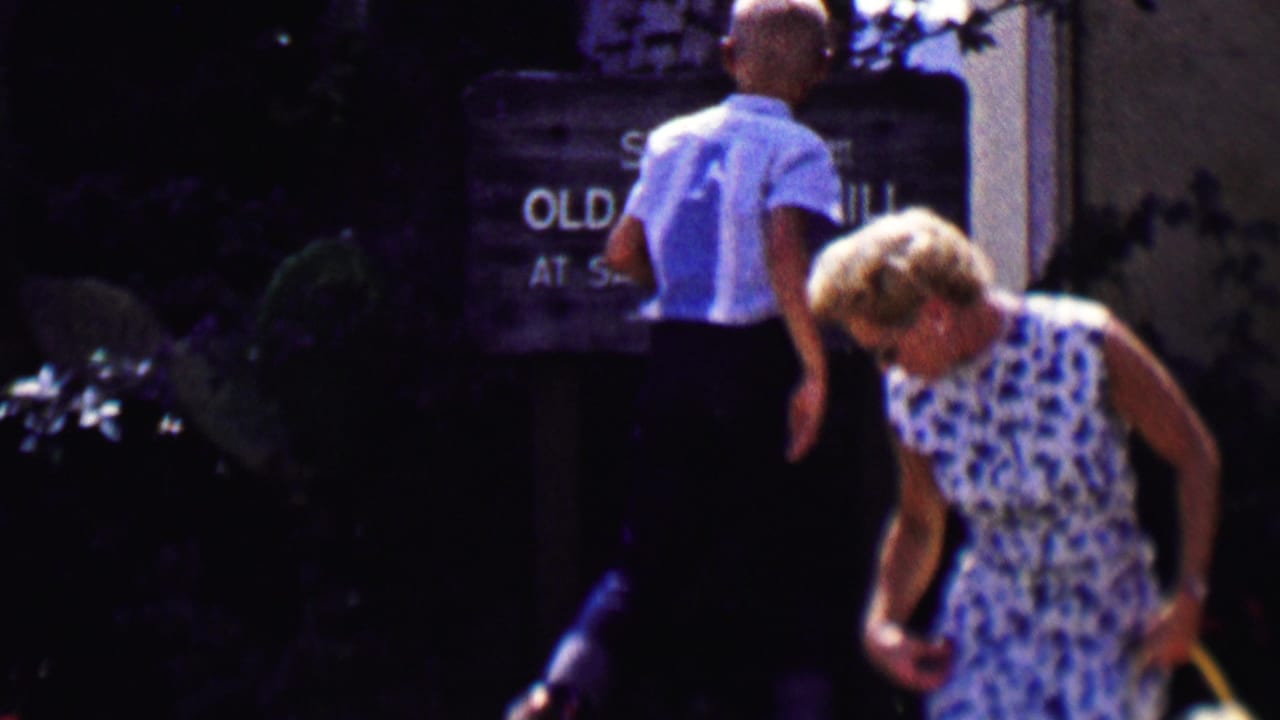 In 1955 Solvang, California, a woman in a blue-and-white patterned dress and sunglasses moves beside Old Olive Mill signage while a younger male figure stands partly turned away behind her.