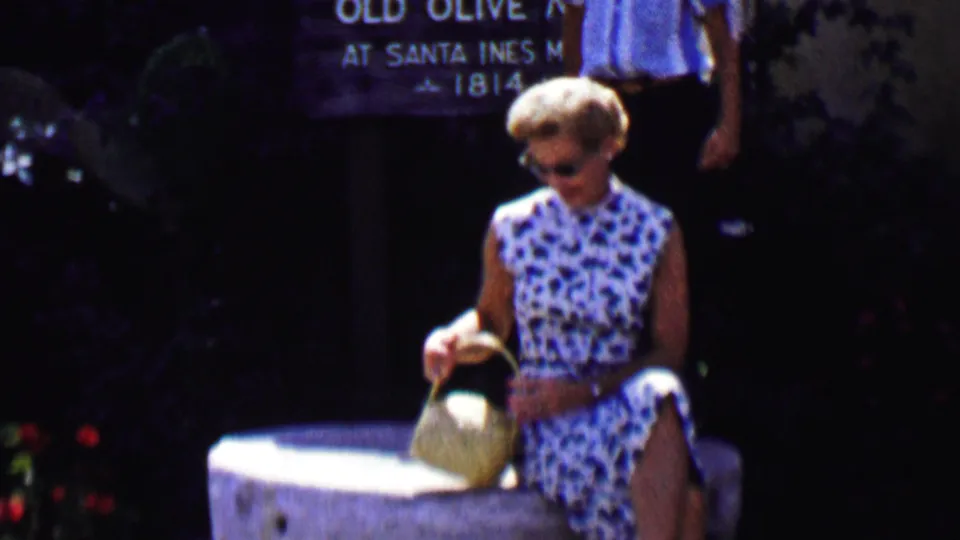 A woman in sunglasses and a sleeveless patterned dress sits on the stone feature with her woven purse in front of Old Olive Mill signage during a sunny 1950s Solvang visit.