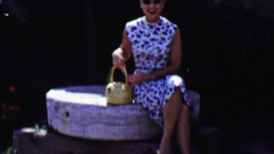 The woman smiles toward the camera while seated on the stone feature, holding a woven purse against the shaded greenery of the Old Olive Mill setting in Solvang.