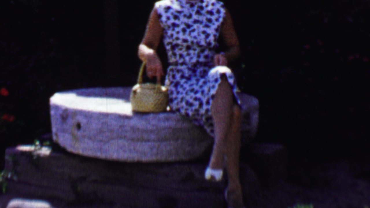 In a 1955 Solvang, California home-movie frame, a woman in a blue and white sleeveless dress sits on a large stone millstone and holds a woven basket against dark foliage.