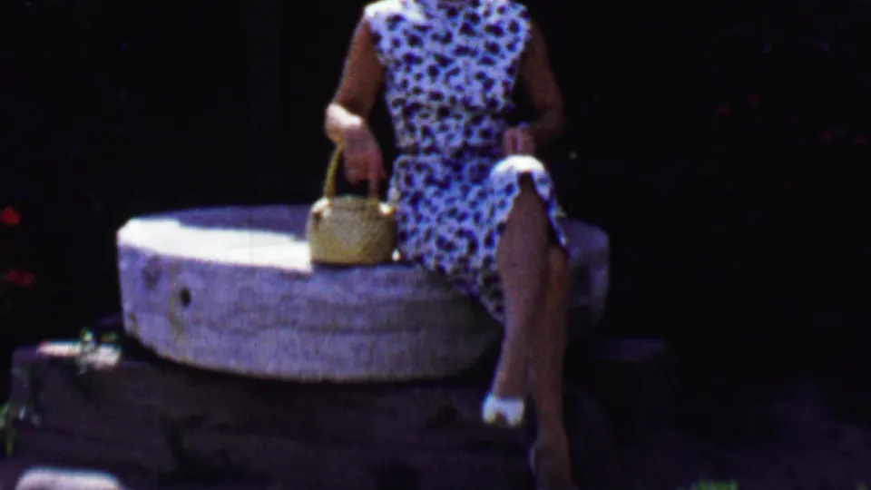 In a 1955 Solvang, California home-movie frame, a woman in a blue and white sleeveless dress sits on a large stone millstone and holds a woven basket against dark foliage.