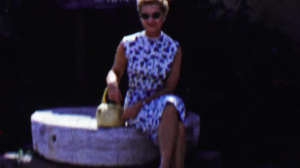 The woman remains seated on the stone millstone in Solvang, wearing sunglasses and a patterned sleeveless dress while holding a small woven basket.