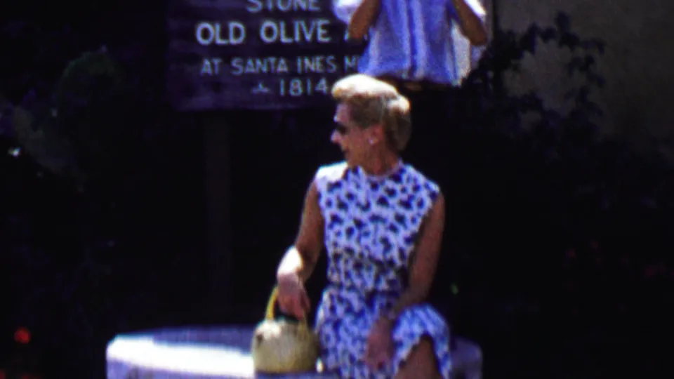 A 1955 Solvang frame shows the woman on the stone millstone with a readable sign behind her that appears to reference an old olive at Santa Ines Mission and the year 1814.
