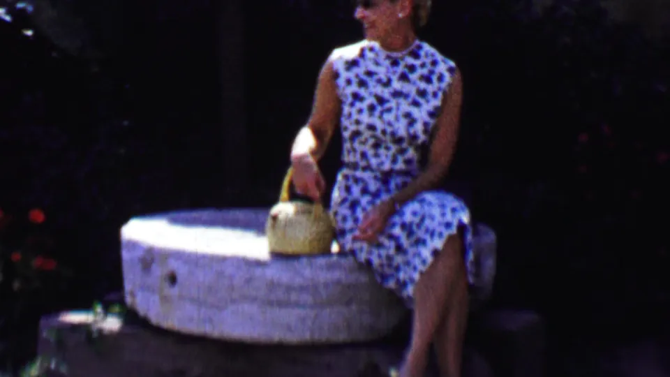 The woman turns her head to the side while seated on the millstone, holding the basket in a shaded garden scene from the Solvang home movie.