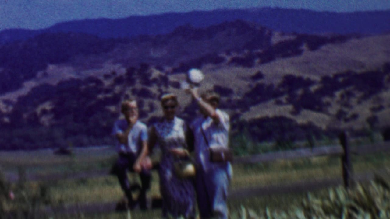 In the supplied 1955 Solvang, California home-movie context, a family group stands near a fence in dry grass with rural hills and mountains in the background.