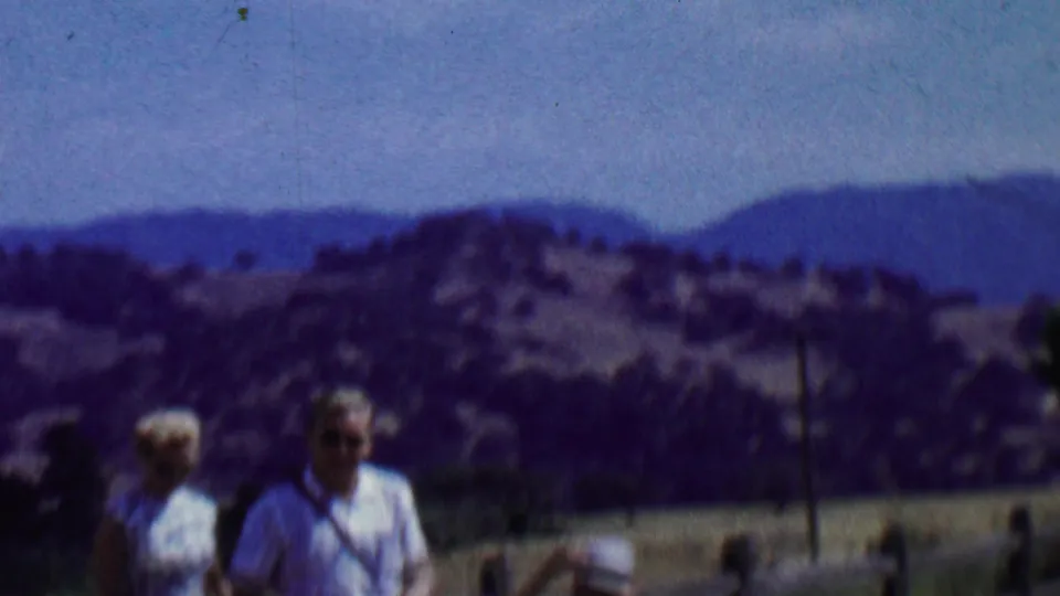Two adults are partially framed at the bottom of a 1950s Solvang home-movie shot, with a fence, open hills, and distant mountains occupying most of the view.