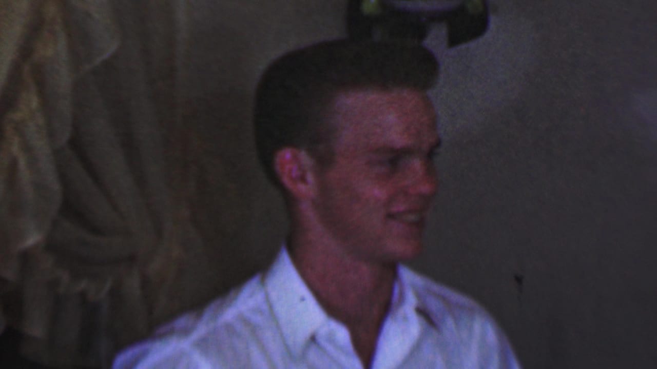In 1963 Terre Haute home-movie footage, a young male in a white collared shirt smiles while seated or standing in a dim indoor setting.