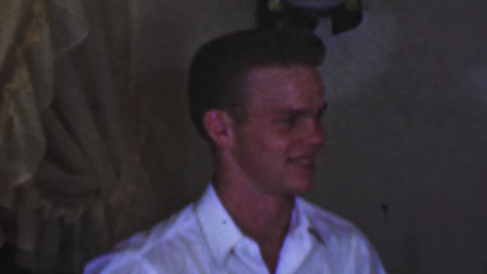 In 1963 Terre Haute home-movie footage, a young male in a white collared shirt smiles while seated or standing in a dim indoor setting.