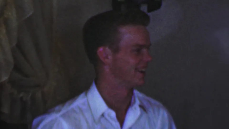 In a dim Terre Haute interior, the young male turns sideways and laughs, with curtains or hanging fabric behind him.