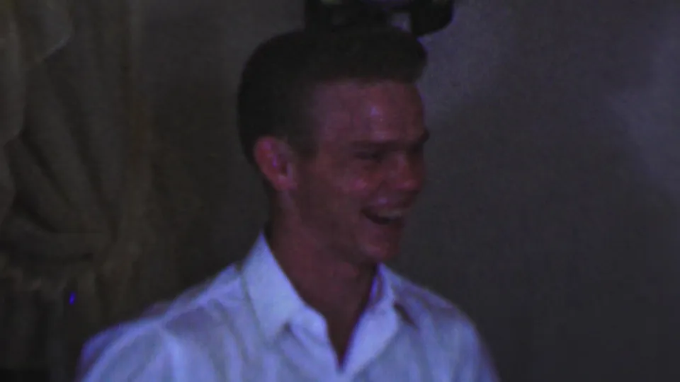 The subject laughs broadly in close framing, with low light, soft focus, and domestic interior details visible behind him.