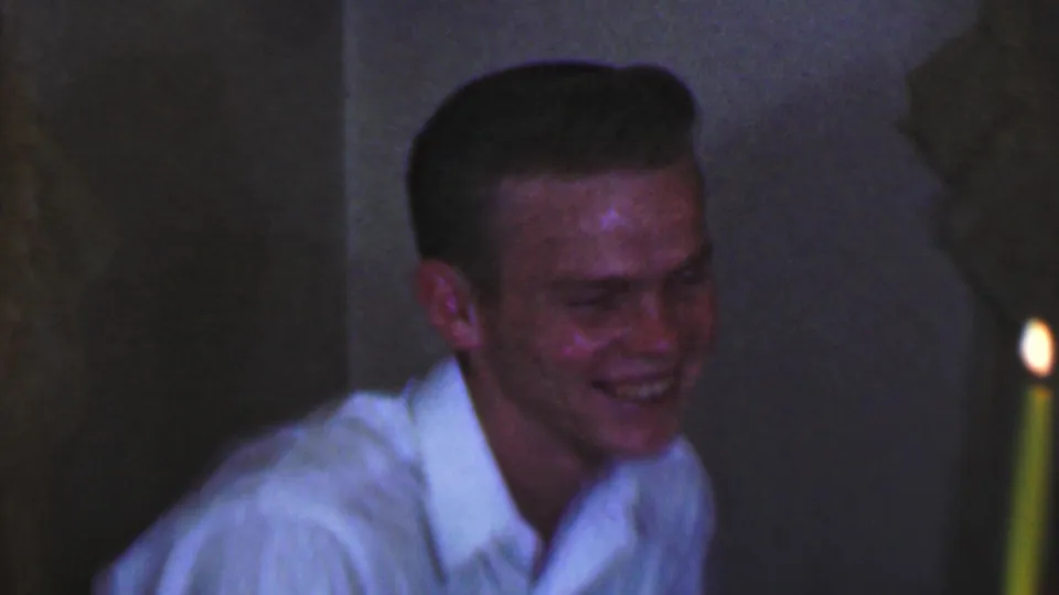The young male leans forward while laughing, with a lit candle at the right edge of the dim 1963 indoor home-movie frame.