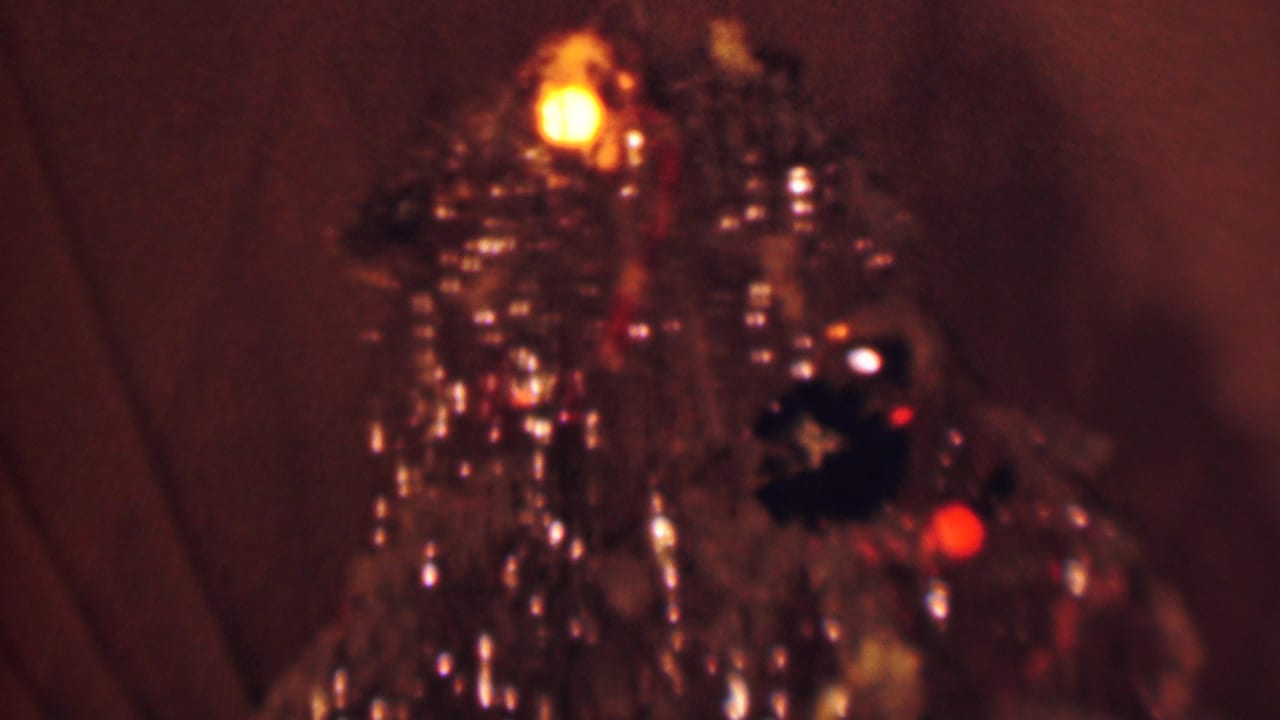 1963 Terre Haute home-movie frame showing a close-up of a decorated Christmas tree with shiny tinsel, warm bulbs, dark ornaments, and curtains in the background.