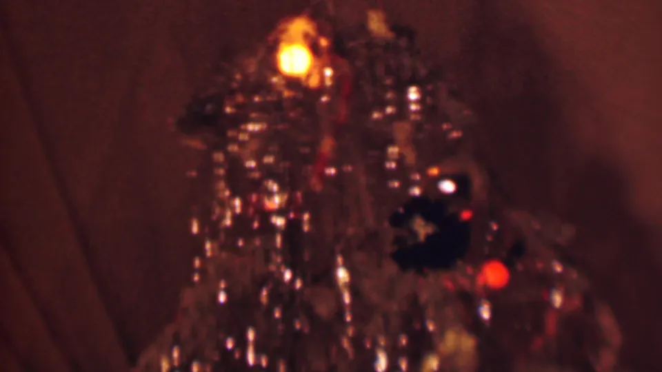 1963 Terre Haute home-movie frame showing a close-up of a decorated Christmas tree with shiny tinsel, warm bulbs, dark ornaments, and curtains in the background.