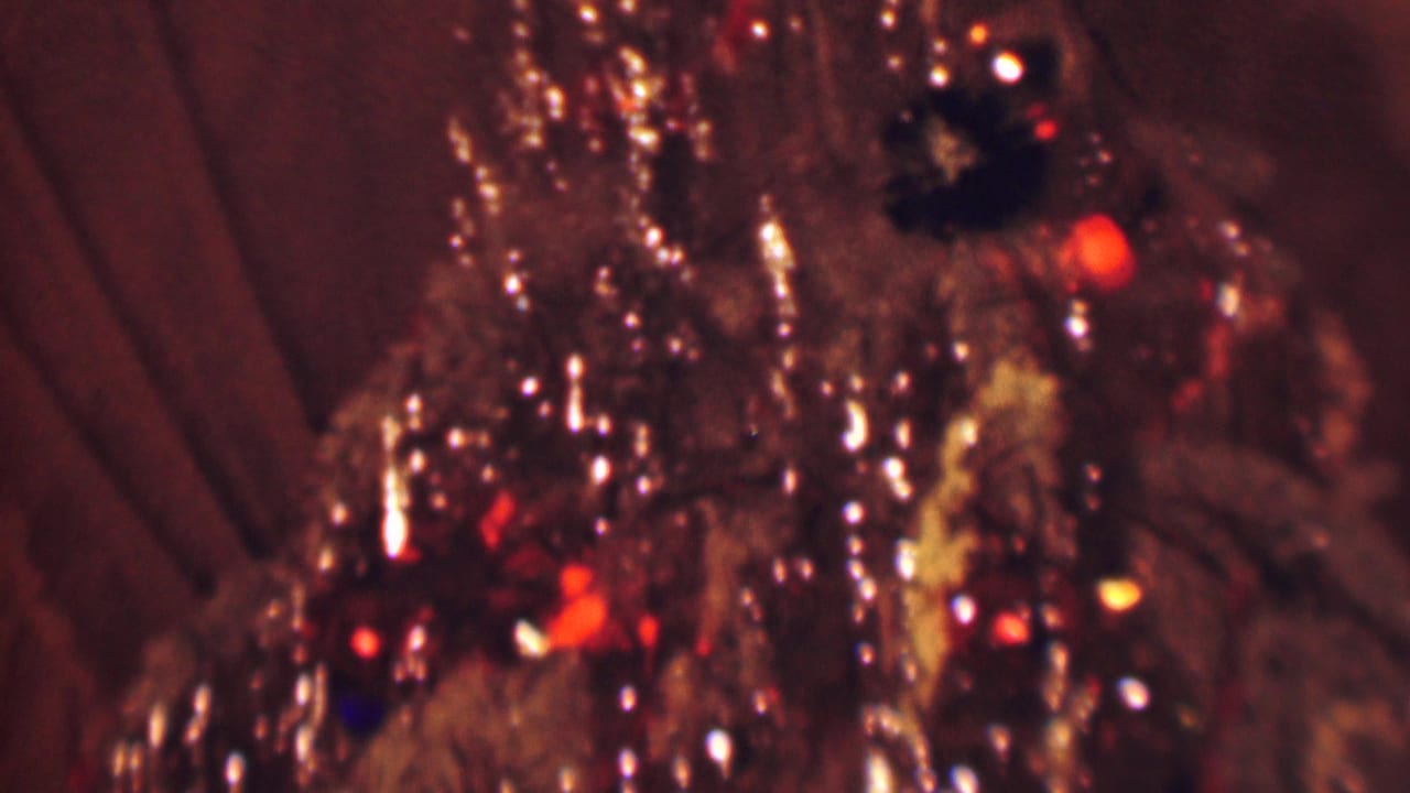 Blurred close-up of Christmas tree tinsel, red and orange lights, and dark ornaments.