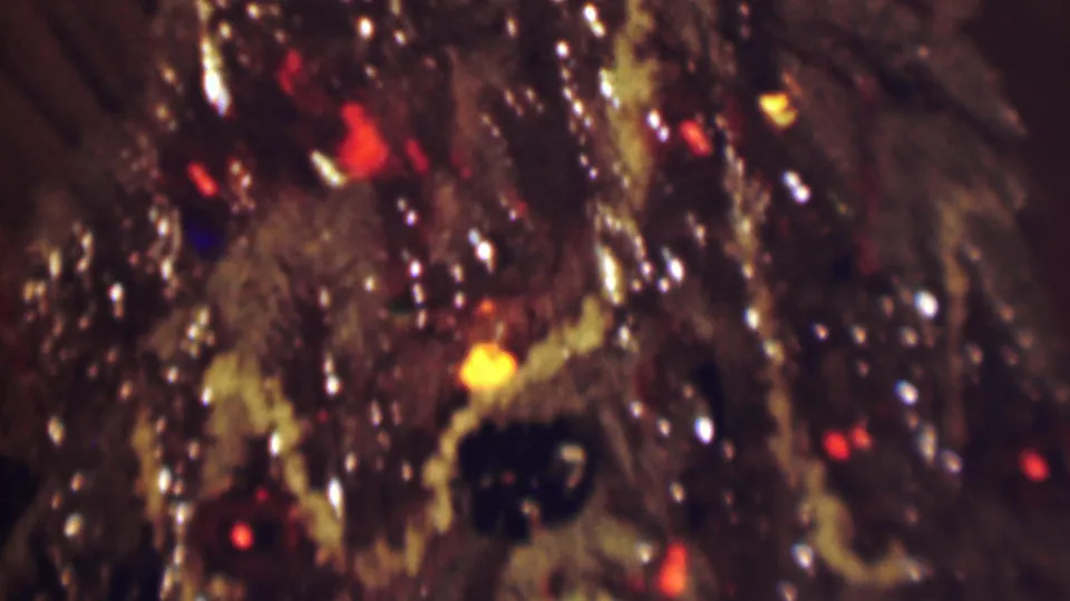 Close-up from the 1963 home movie showing flocked evergreen branches, a dark round ornament, and glowing holiday lights rendered with soft 8mm grain.