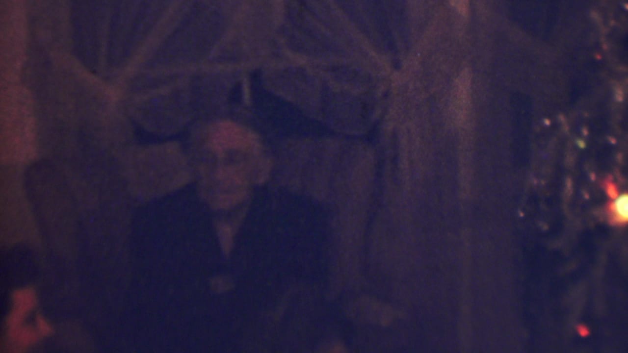 The older woman remains seated in the low-lit Terre Haute Christmas gathering, facing toward the camera with holiday tree lights visible at the right edge.