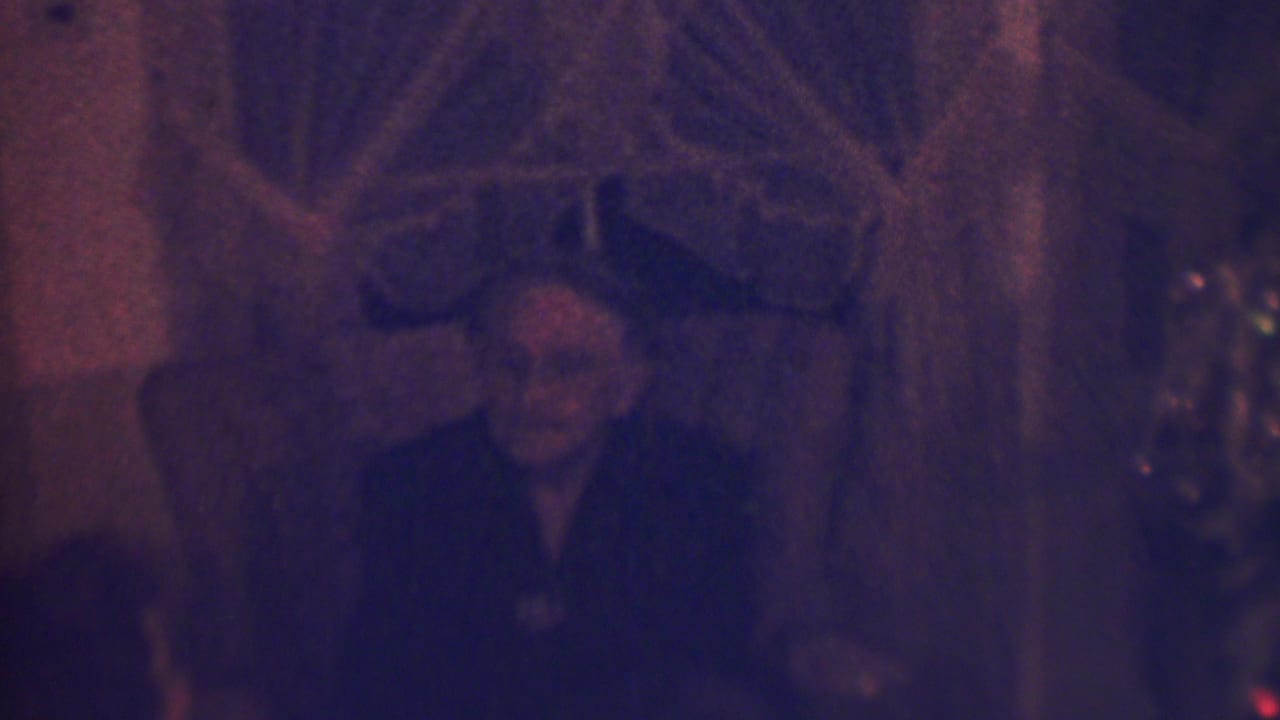 The seated older woman appears to smile or react during the 1963 home-movie Christmas scene, with the tree and domestic interior partly visible around her.