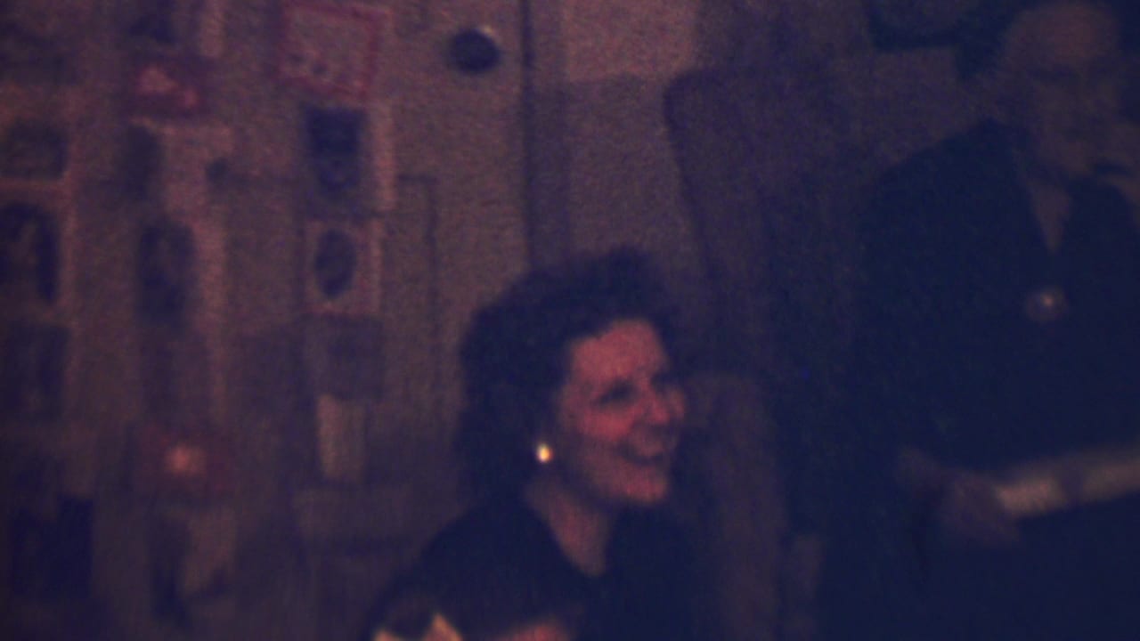 In a 1963 Terre Haute, Indiana home-movie frame, a woman with dark styled hair and earrings laughs while seated in a dim holiday interior, with wall cards or photographs behind her.