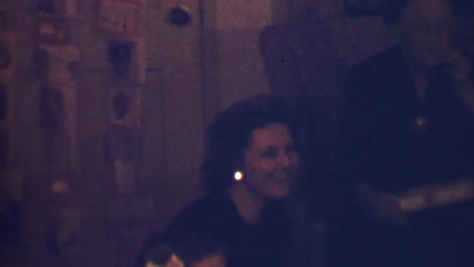 A low-light 8mm frame from the Terre Haute home gathering shows the seated woman smiling while a standing man appears partly obscured behind her near the wall display.