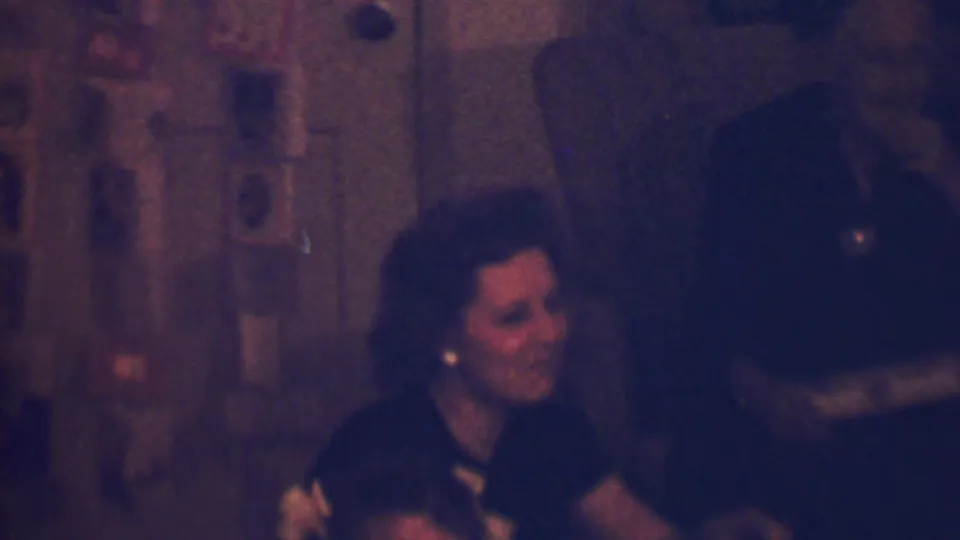 The final frame shows the woman still smiling in the dim 1963 Terre Haute family interior, with wall cards or photographs and a darkened area of the room in the background.