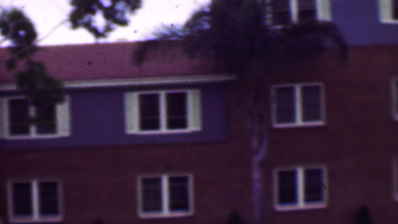1961 San Diego home-movie frame showing the upper levels of a multi-story apartment building with white-trimmed windows, a red-tiled roof, and tree branches in the foreground.