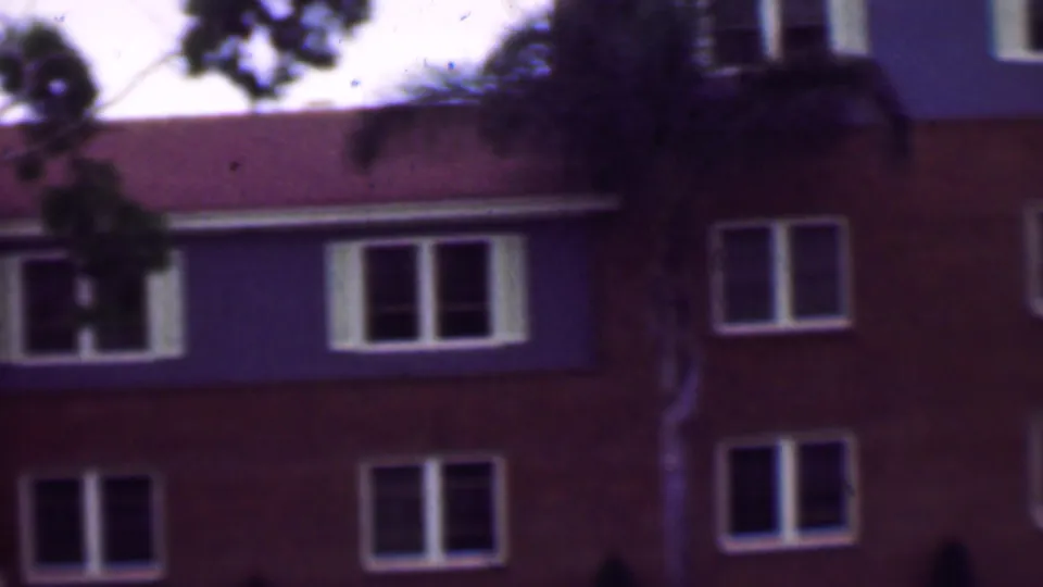 1961 San Diego home-movie frame showing the upper levels of a multi-story apartment building with white-trimmed windows, a red-tiled roof, and tree branches in the foreground.