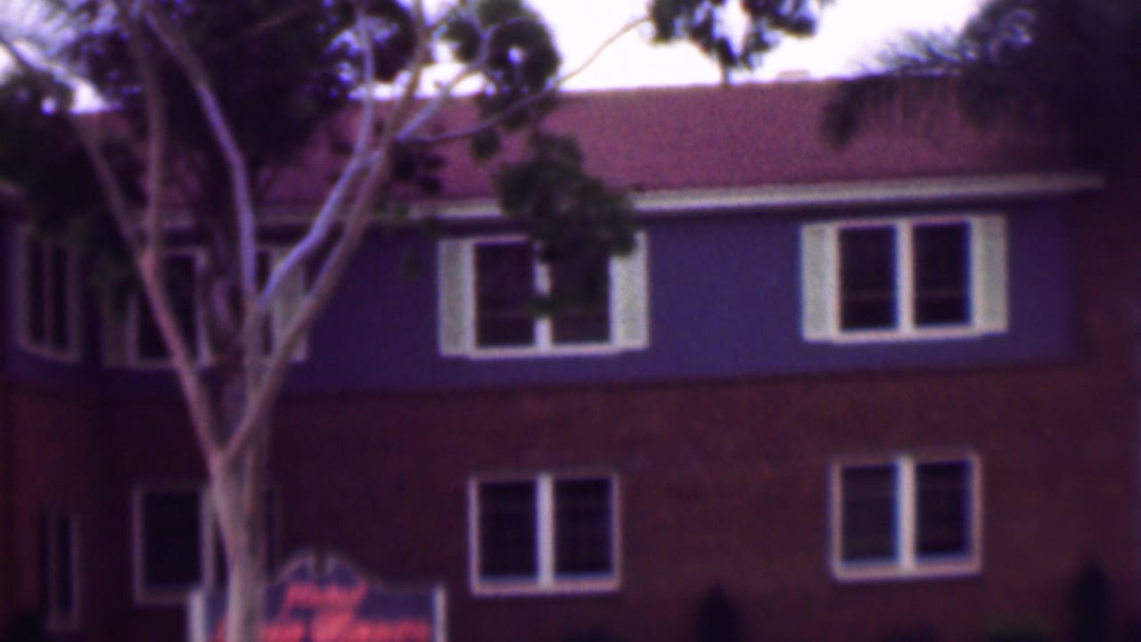 1961 San Diego frame showing a brown and purple-blue apartment exterior with white-framed windows, palm foliage, and a red vehicle passing low in the foreground.