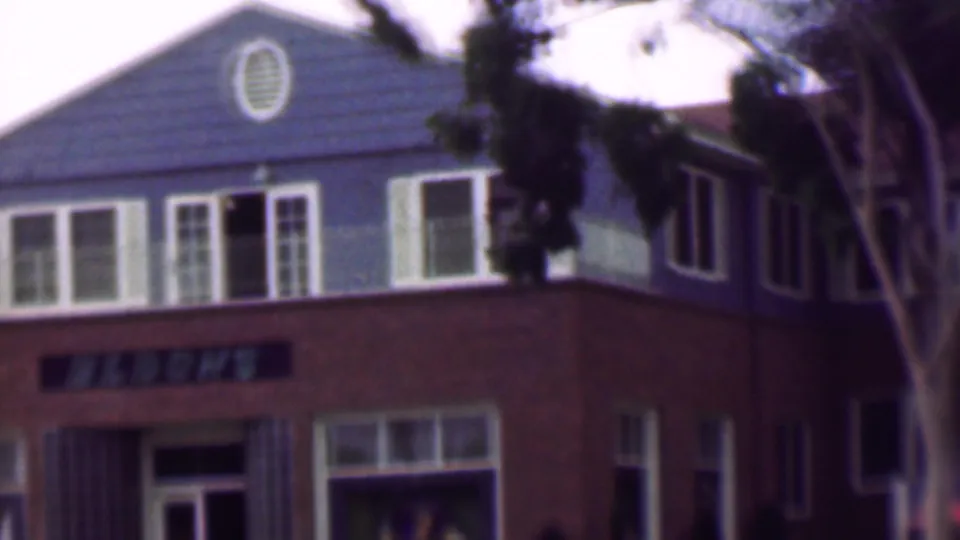 1961 San Diego frame showing the front portion of a multi-story apartment building with a gabled blue-gray upper facade, white-trimmed windows, brown lower wall, entrance area, and trees at the edges.