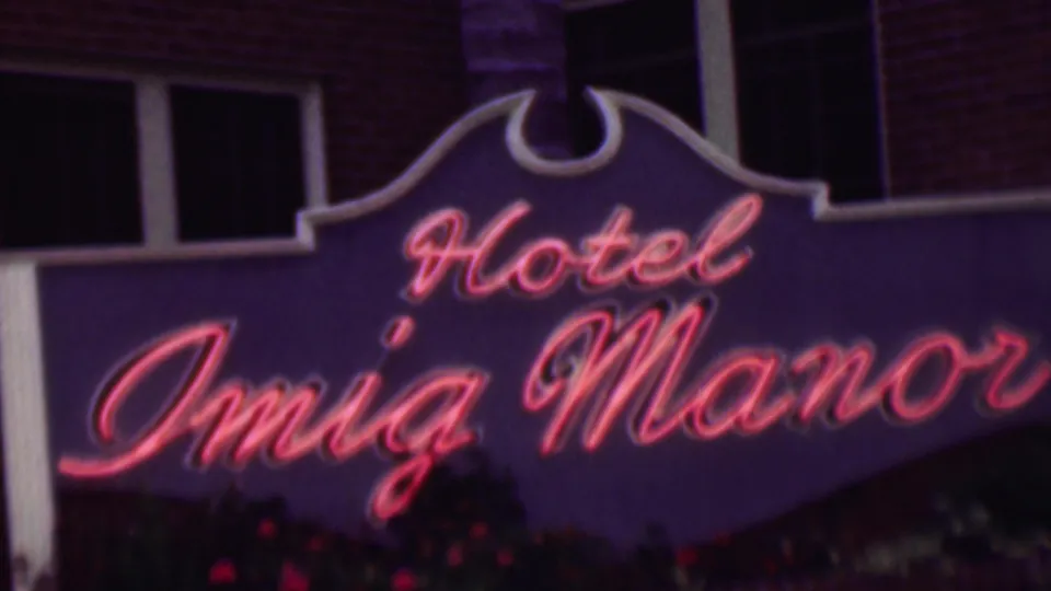 1961 San Diego home-movie frame of the Hotel Imig Manor neon sign, with red script lettering, purple signboard, brick facade, and small flowers below.