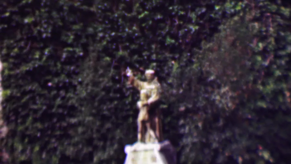 1961 San Diego 8mm frame showing a pale male statue with one arm raised or pointing, set on a pedestal before dense green ivy.