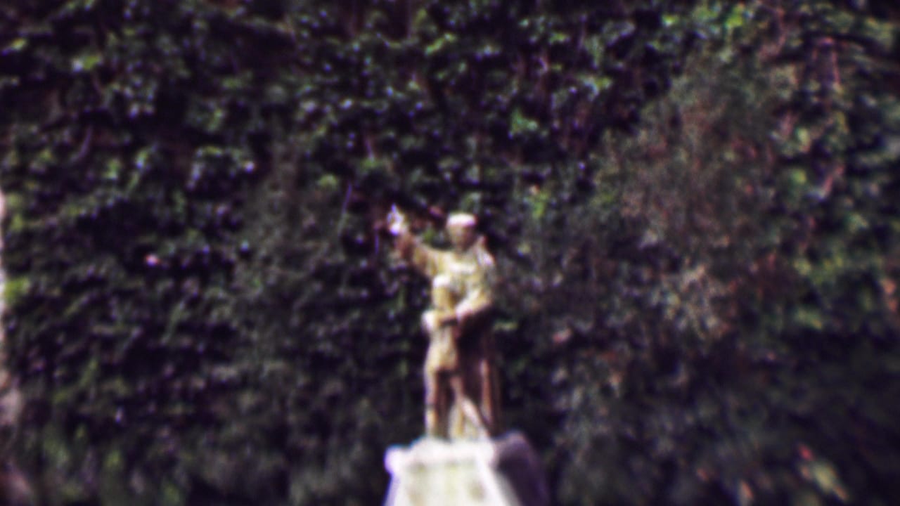 1961 San Diego 8mm frame showing a weathered stone statue of a man waving while holding a boy figure, set before a dense ivy-covered wall.