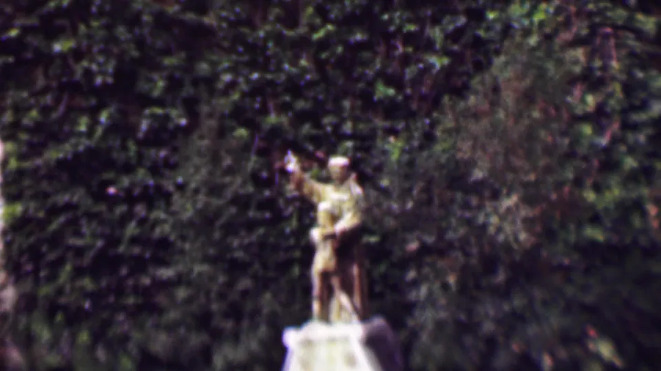 1961 San Diego 8mm frame showing a weathered stone statue of a man waving while holding a boy figure, set before a dense ivy-covered wall.