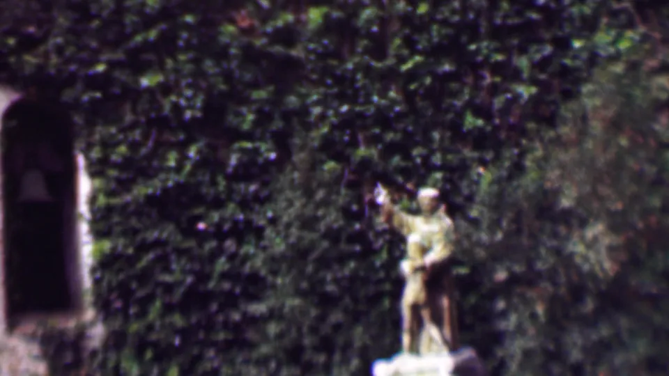 1961 San Diego 8mm frame showing the statue beside arched stone openings, with ivy and shrubs filling much of the background.