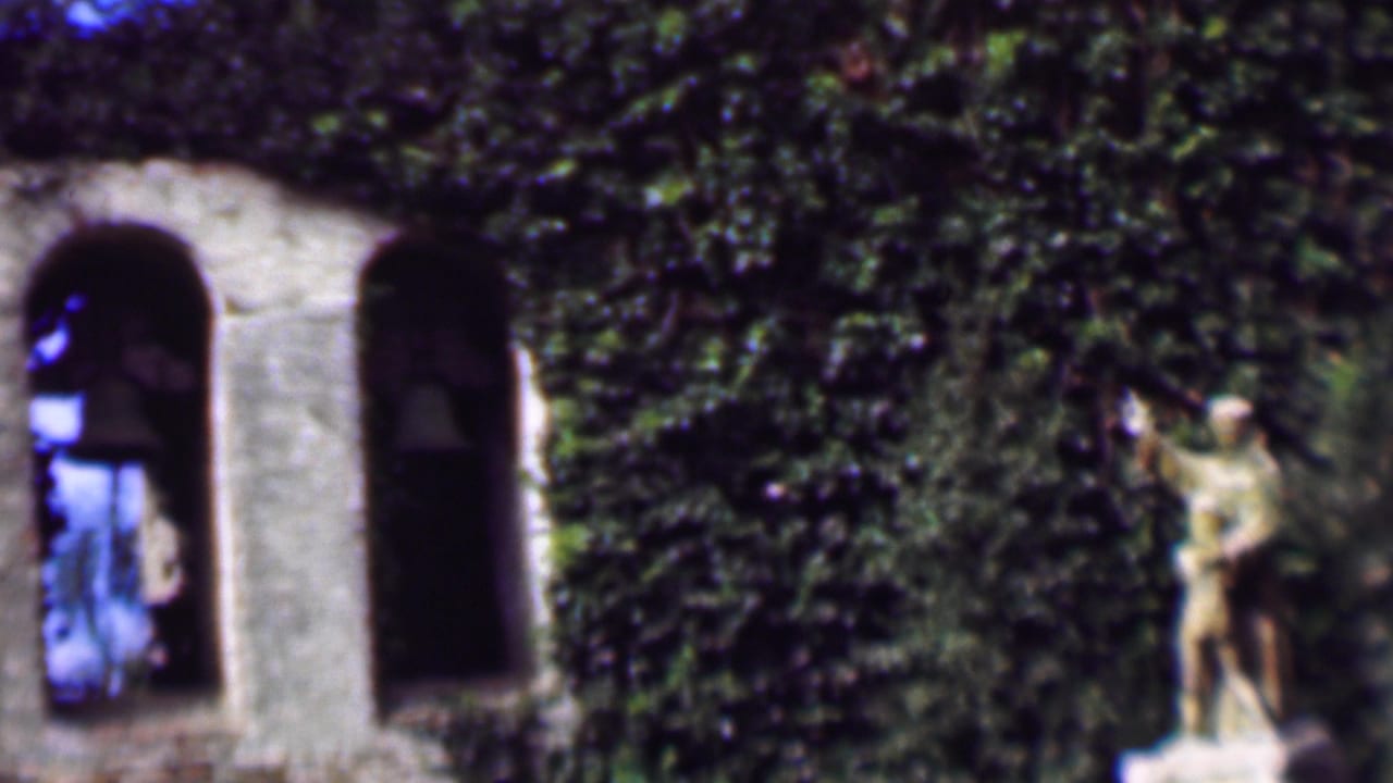 1961 San Diego home-movie frame showing an ivy-covered stone bell tower with arched openings, hanging bells, and a small statue near the center.