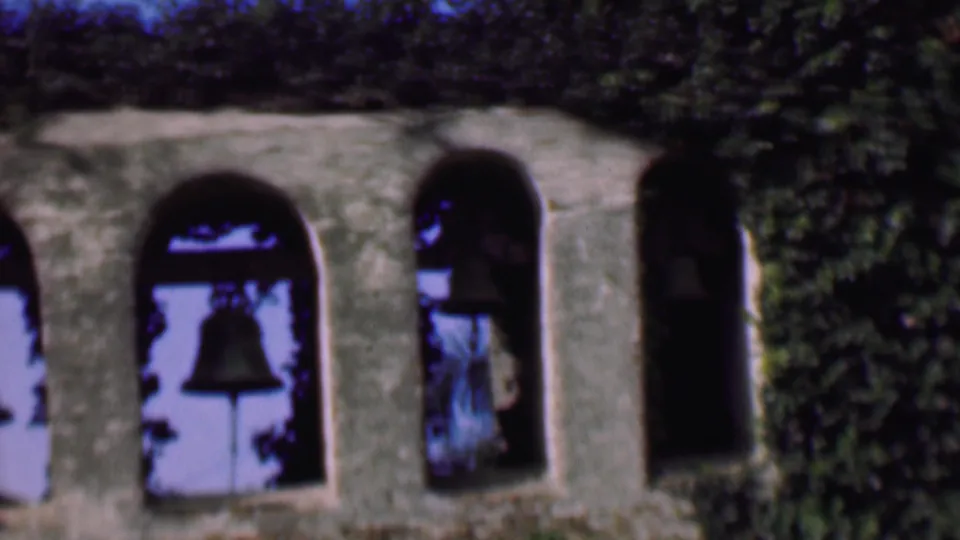 1961 San Diego home-movie frame showing a closer section of the stone bell tower, with arched openings, bells in shadow, and ivy climbing between the masonry.