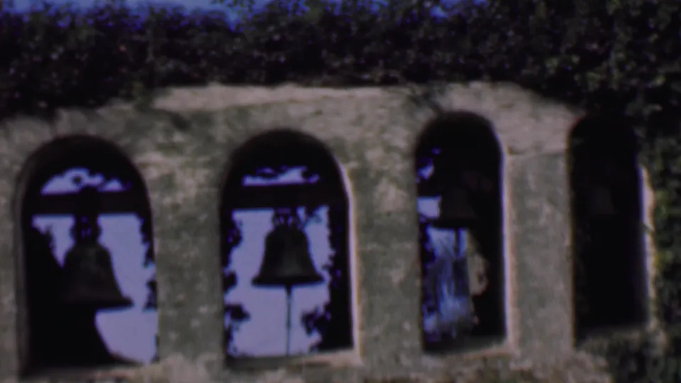 1961 San Diego frame showing several stone arch openings with suspended bells, heavy ivy along the top, and high-contrast home-movie exposure.