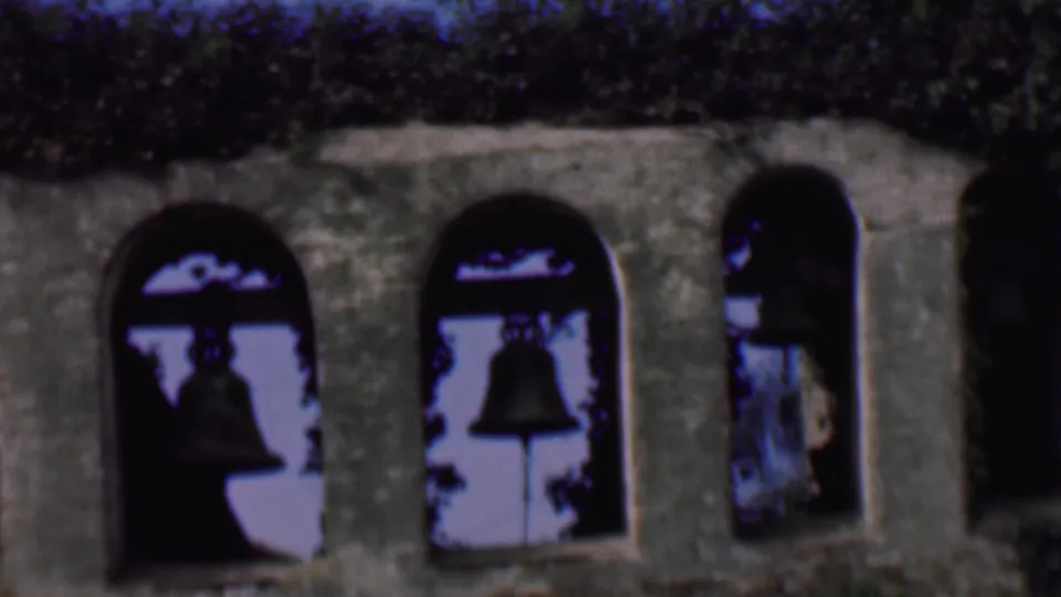 1961 San Diego frame of an old stone bell tower facade, with several arched bays, dark bells, ivy, and soft blue sky behind the openings.