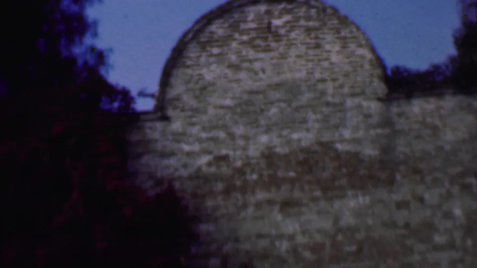 1961 San Diego frame showing the rounded upper stonework of the bell-tower structure, bordered by dark vegetation and pale sky.