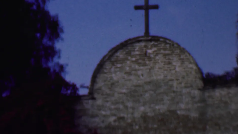 1961 San Diego frame with a dark cross rising above a rounded stone church dome, bordered by tree silhouettes and blue sky.
