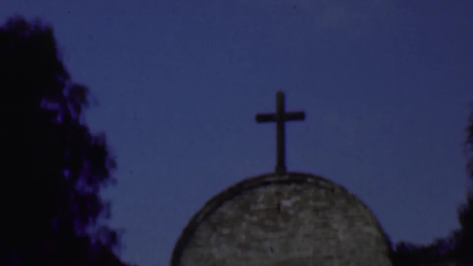 1961 San Diego frame isolating the cross and upper stone dome against a broad blue sky with tree silhouettes at the edges.