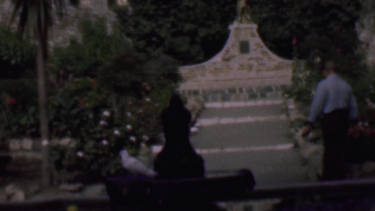 In 1961 San Diego home-movie footage, a man in a light shirt and dark trousers walks away from the camera along a lush garden path toward a stone bird bath or fountain.