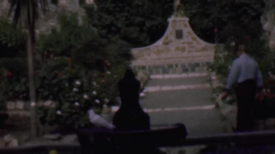 In 1961 San Diego home-movie footage, a man in a light shirt and dark trousers walks away from the camera along a lush garden path toward a stone bird bath or fountain.