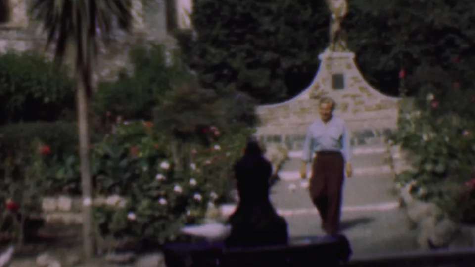 A man in a light shirt and dark trousers walks along a paved garden path in San Diego, with flowers, tall foliage, and a pale stone bird bath or fountain behind him.