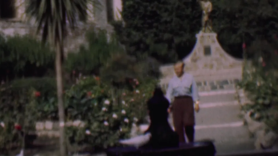 The man moves closer to the foreground beside a dark bench, with a stone garden feature, flowering beds, and tall foliage visible in the soft 8mm frame.