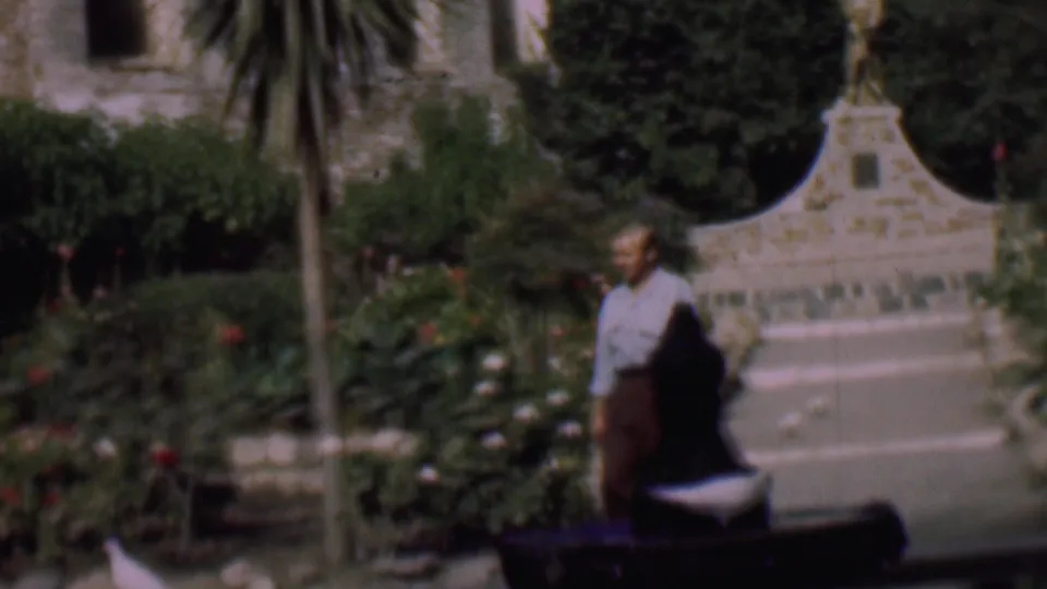 A man passes near the dark foreground bench in a planted San Diego garden, with a pale stone bird bath or fountain and decorative arched stonework behind him.