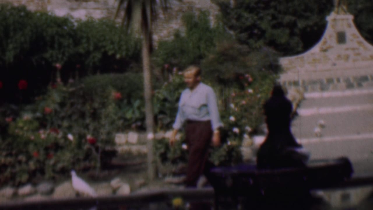 In 1961 San Diego home-movie footage, a man in a light shirt and brown trousers walks through a rose garden beside a pond where white pigeons gather.
