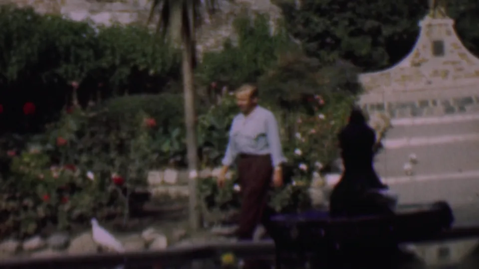 In 1961 San Diego home-movie footage, a man in a light shirt and brown trousers walks through a rose garden beside a pond where white pigeons gather.